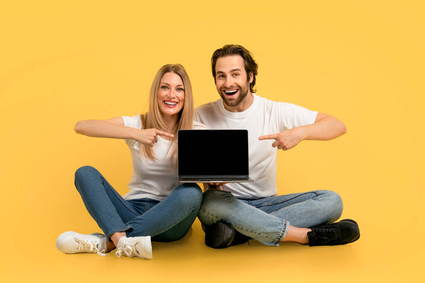 Happy, beyaz tişörtlü beyaz kadın ve adamdan ilham alarak ekranı boş ekranla gösteren sarı arka planda, stüdyoda izole bir şekilde oturuyor. Harika reklam ve teklif, web sitesi, online uygulama - Fotoğraf, Görsel