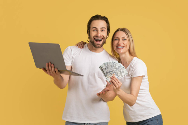 Веселий молодий чоловік і дружина в білих футболках з ноутбуком і багато доларів радіють онлайн перемозі, ізольовані на жовтому тлі. Велике оголошення і пропозиція, людські емоції, фінанси і гроші. - Фото, зображення
