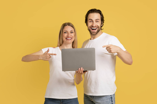Glad young caucasian wife and husband in white t-shirts show fingers at laptop, isolated on yellow background, studio. Social networks and blog recommendation, ad and offer people emotions, great app - Photo, Image