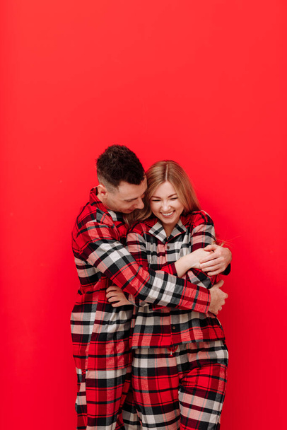 Happy Saint Valentine's Day. Young couple in the same pyjamas is kissing and hugging on red background.Man and woman in love are enjoying spending time together while celebrating, copy space for text. - Фото, зображення