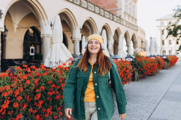 Young happy woman traveler in yellow hat listening to music through wireless earphones on urban background. Stylish female foreigner examines architectural monument during her vacation - Photo, Image