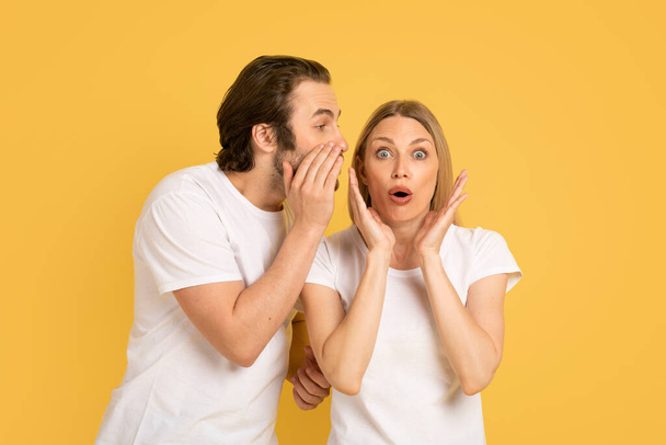 Cheerful young caucasian guy whispering into ear of shocked woman in white t-shirt, isolated on yellow background, studio. Human emotions, gossip, news, great ad and offer, love and relationship - Photo, Image