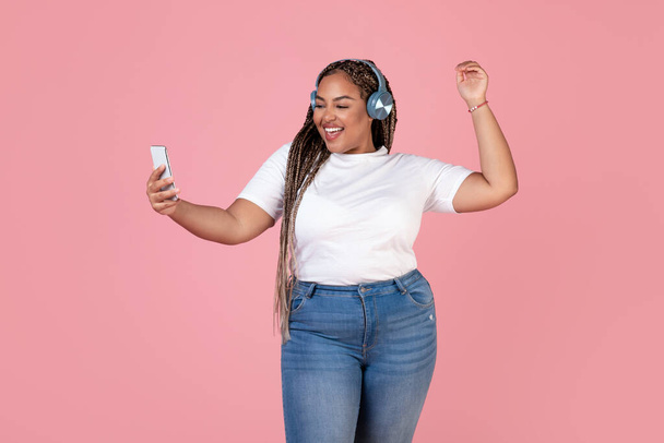 Musical App. Joyful African American Oversized Lady Listening To Music Holding Smartphone Wearing Earphones, Enjoying Favorite Song Dancing In Studio Over Pink Background - Photo, Image