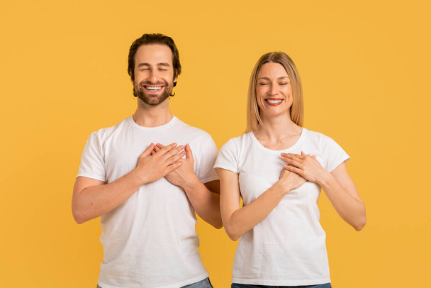 Glad young caucasian lady and male in white t-shirts make gratitude gesture, press hands to chest, isolated on yellow background, studio, empty space. Hope, wish, ad and offer, love, relationship - Photo, Image