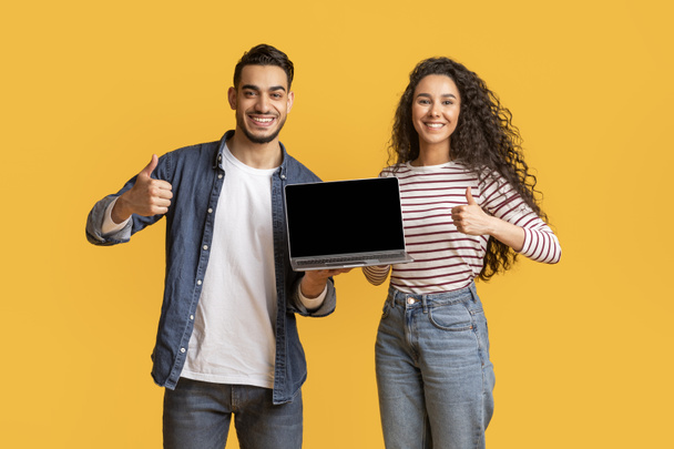 Sonriendo joven pareja árabe que sostiene el ordenador portátil con pantalla negra y mostrando pulgares hacia arriba, hombre y mujer de Oriente Medio feliz recomendando un nuevo sitio web, de pie aislado sobre fondo amarillo, burla - Foto, Imagen