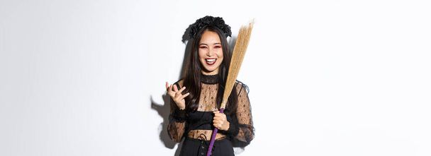 Image of witch in gothic lace dress and black wreath making evil laugh and holding broom, celebrating halloween, standing over white background. - Photo, Image