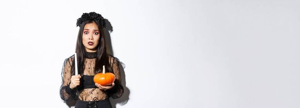 Asyalı bir kadının paniklemesi cadılar bayramında büyücülükten korkması, cadı kostümünün içinde mum ve balkabağıyla durması.. - Fotoğraf, Görsel