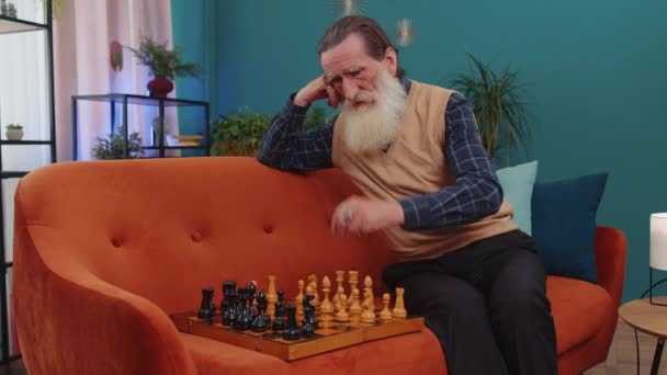 Εστιασμένη ανώτερος παππούς άνθρωπος chessman κάθεται στο σπίτι καναπέ αίθουσα μάθησης παίζοντας σκάκι επιτραπέζιο παιχνίδι και μόνο, εκπαιδευτική οικιακή δραστηριότητα αναψυχής, στρατηγικές δεξιότητες. Ηλικιωμένοι συνταξιούχος παππούς συνταξιούχος - Πλάνα, βίντεο