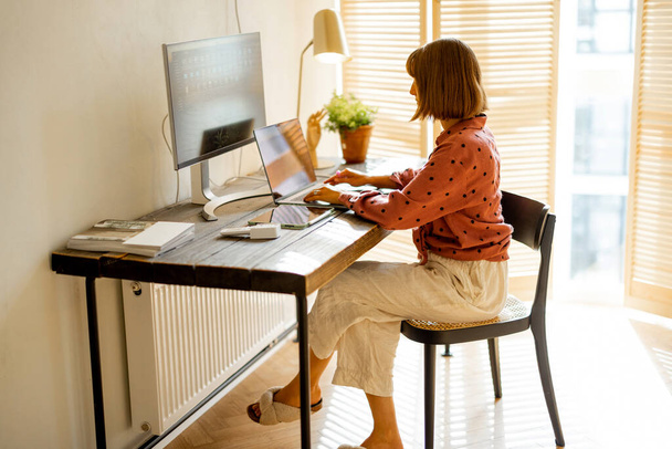 Γυναίκα εργάζεται σε υπολογιστές, ενώ κάθεται από ξύλινο τραπέζι στο ηλιόλουστο δωμάτιο στο σπίτι. Έννοια της απομακρυσμένης εργασίας από το σπίτι σε ζεστή ατμόσφαιρα - Φωτογραφία, εικόνα
