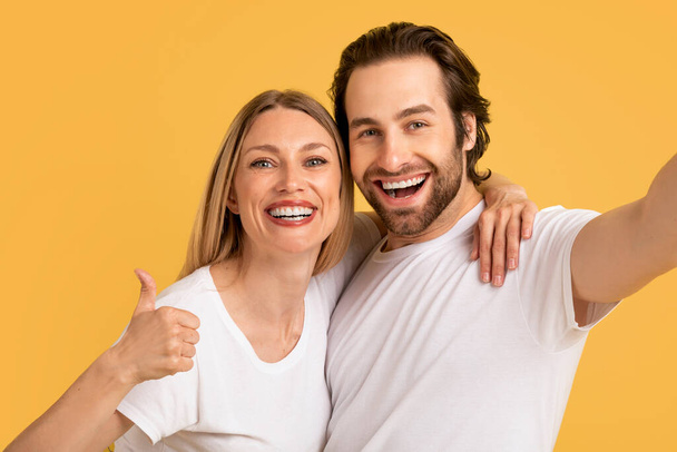 Portret van vrolijke jonge blanke echtparen in witte t-shirts tonen duim omhoog, selfie nemen, geïsoleerd op gele achtergrond, close-up. Foto voor sociale netwerken, blog, reclame en aanbod, emoties, videogesprek - Foto, afbeelding