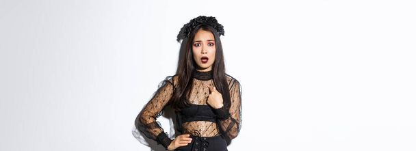 Beleidigte und verwirrte asiatische Frau im Halloween-Kostüm zeigt auf sich selbst, trägt schwarzes Kleid der bösen Hexe auf Party, steht über weißem Hintergrund. - Foto, Bild