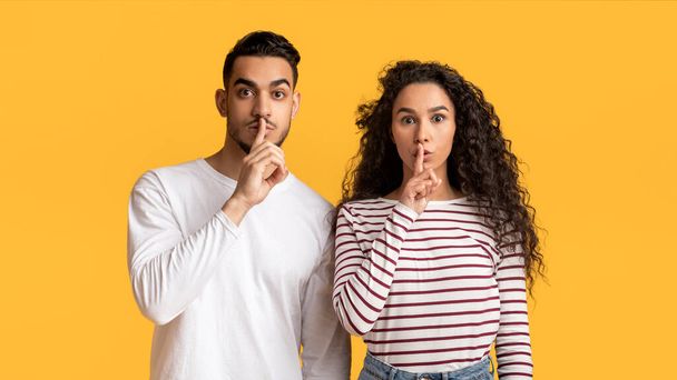 Junge arabische Männer und Frauen zeigen shh Geste in die Kamera, geheimnisvolles Paar aus dem Nahen Osten hält Zeigefinger auf den Lippen, macht Schweigezeichen, während sie zusammen vor gelbem Hintergrund stehen, kopieren Raum - Foto, Bild