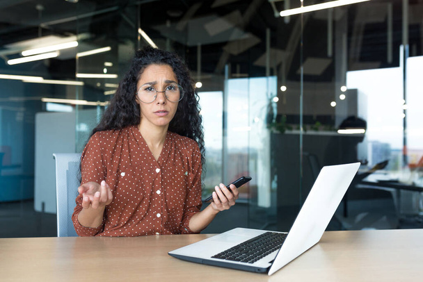 Frustrated and sad business woman with phone in hands looking at camera, latin american woman working with laptop inside modern office building, unclear emotional state of female employee. - Photo, Image