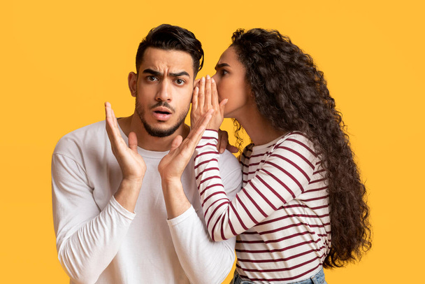 Young Arab Woman Sharing Gossips With Her Shocked Boyfriend, Whispering To His Ear, Millennial Middle Eastern Female Telling Secret To Spouse While They Standing Together Over Yellow Background - Photo, Image