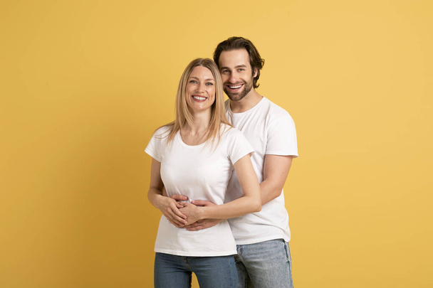 Love, family relationship and romance. Smiling young caucasian male hugging woman in white t-shirts, enjoy time together, isolated on yellow background, studio. Sale, ad and offer, people emotions - Foto, Imagem