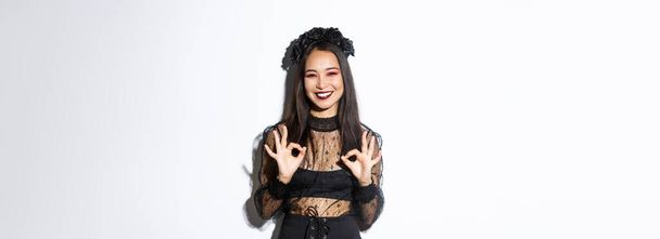 Веселая красивая азиатская женщина в ведьминском платье показывает нормальные жесты и улыбаясь удовлетворены, одобрить Хэллоуин костюм или рекламу, стоя на белом фоне. - Фото, изображение