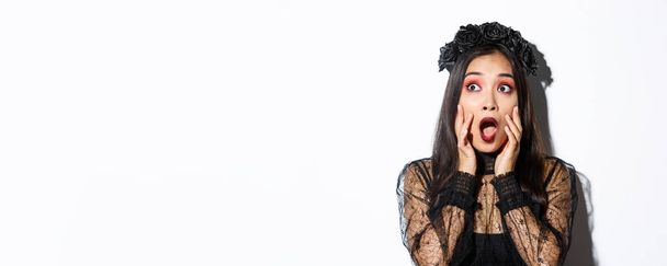 Close-up of scared asian woman in halloween costume of witch looking frightened at upper left corner, gasping and standing ambushed over white background. - Foto, afbeelding