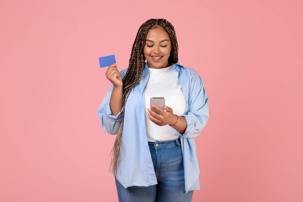Compras móviles. Mujer afroamericana alegre usando tarjeta de crédito y teléfono celular haciendo compras de pago más ropa de tamaño de pie sobre fondo rosa. Tiro de estudio - Foto, imagen