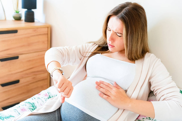 Pregnant contractions time. Pregnancy woman watching clock, holding baby belly. Childbirth time, contractions pain. Concept of pregnant, maternity, expectation for baby birth - 写真・画像