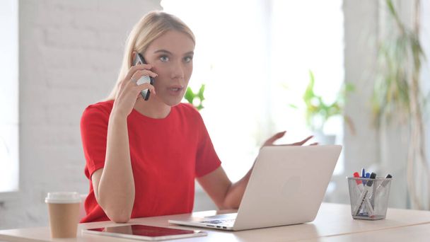 Злая молодая женщина разговаривает по телефону во время использования ноутбука в офисе - Фото, изображение