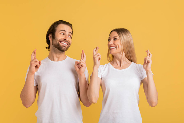 Smiling young caucasian female and male in white t-shirts make wishes and cross fingers, isolated on yellow background, studio. Emotions, hope, dream and success, great ad and offer and gestures - Φωτογραφία, εικόνα