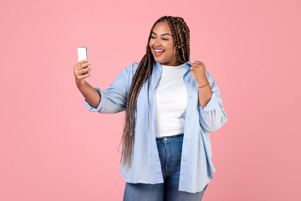 ハッピーアフリカ系アメリカ人レディービデオピンクスタジオの背景にスマートフォンを掲出保持を呼び出します。女性コミュニケーションと電話でセルフィーを作る楽しさを持っています.テクノロジーとモバイル通信 - 写真・画像