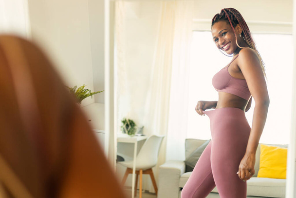 Portrait of happy african american young woman looking herself reflection in mirror at home and enjoying result of successful slimming, copy space - Photo, image