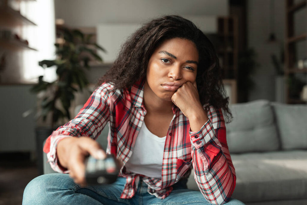entediado triste muito millennial afro-americano feminino com controle remoto, assistir TV notícias ou filme, sentado no sofá na sala de estar interior, espaço livre. Tempo de lazer, ociosidade em casa e emoções das pessoas - Foto, Imagem