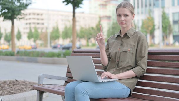 Отклонение молодой женщины в отрицании во время использования ноутбука сидит на открытом воздухе на скамейке - Фото, изображение