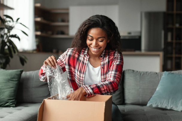 Тысячелетняя африканка открывает картонную коробку с покупками и распаковывает посылку, наслаждаясь продажей в интерьере гостиной. Доставка и онлайн шоппинг, новые нормальные и человеческие эмоции - Фото, изображение