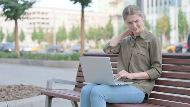 Молодая женщина с болями в шее с помощью ноутбука, сидя на открытом воздухе на скамейке - Фото, изображение
