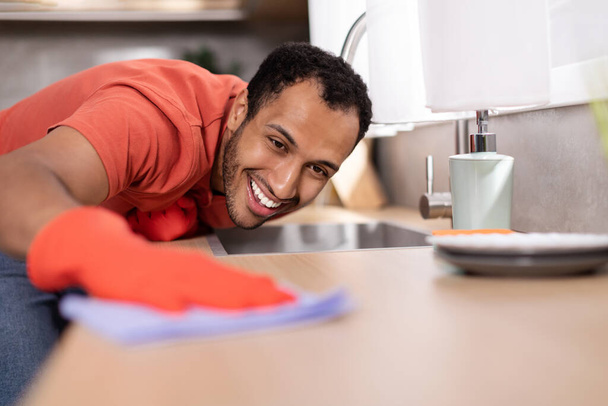 Smiling millennial black male in rubber gloves wipes dust from wooden table in kitchen interior, free space. Housework, cleaning, lifestyle, perfectionism and hygiene, household chores during covid-19 - Photo, Image