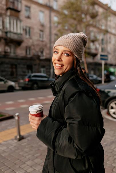 Junges Mädchen mit rotem glücklichem Lächeln lacht und setzt Strickmütze auf. Frau in schickem Mantel und Jeans spaziert mit Kaffee durch Herbststadt - Foto, Bild