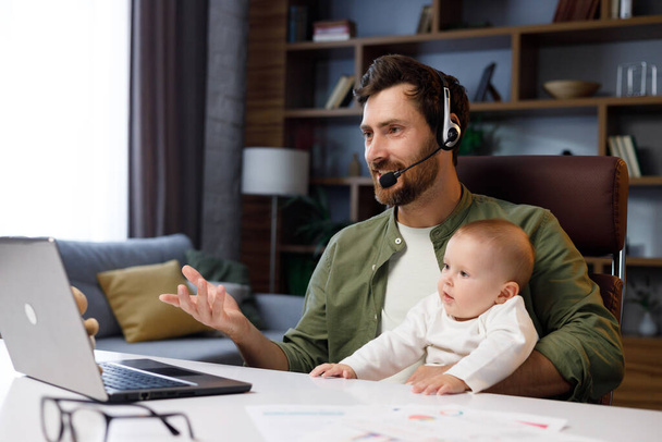 Father on maternity leave, holding an online conference, a meeting in the home office with a small child in his arms. Dad with a child. Freelancer combines child care and work. Paternity leave. - Фото, изображение