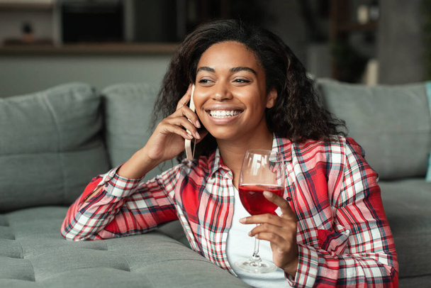 Boldog millenniumi afro-amerikai nő pohárral bor beszél okostelefonon a nappali belsejében. Pihenés és pihenés, dátum és évforduló távolról otthon, ünnep alatt covid-19 - Fotó, kép