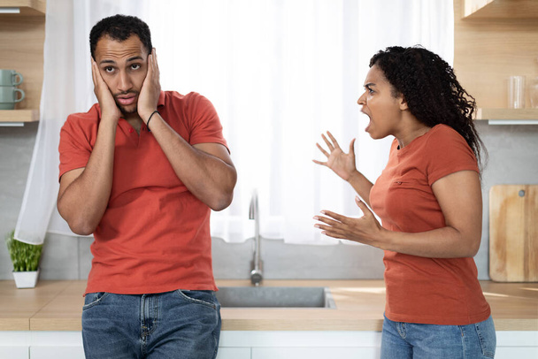 Angry evil aggressive young black wife scolding at offended husband, suffering from pressure and stress in kitchen interior. Human emotions, quarrel, divorce and scandal relationship problems at home - Zdjęcie, obraz