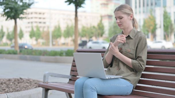 Молодая женщина с болями в запястье с помощью ноутбука, сидя на открытом воздухе на скамейке - Фото, изображение