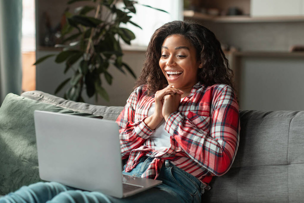 Feliz sorprendido joven afroamericano femenino con portátil se regocija por la victoria y el éxito de las compras en línea en el sofá en el interior de la sala de estar. Gran venta, gran anuncio y oferta, emociones de la gente, grandes noticias - Foto, Imagen