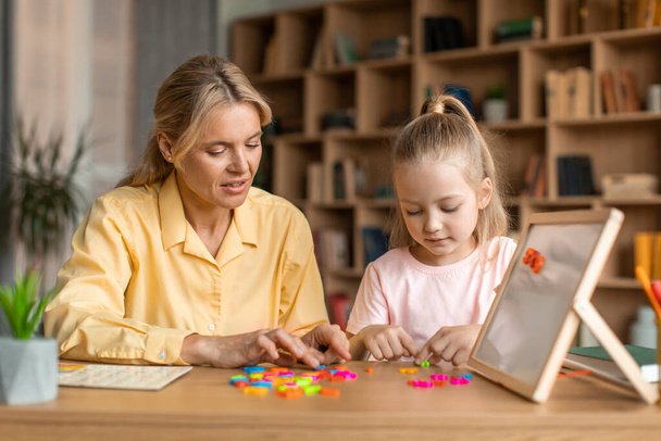 Przyjazna kobieta uczy ładną dziewczynkę alfabetu, specjalista pracujący z dzieckiem, tworząc słowa z kolorowych plastikowych liter razem na zajęciach rozwoju dziecka - Zdjęcie, obraz