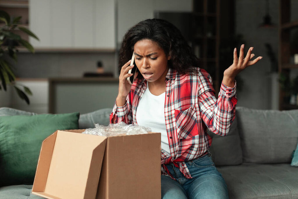 Sad frustrated millennial african american woman opening cardboard box, talking on phone, swearing about mistake with parcel in living room interior. Delivery and online shopping problems, bad service - Foto, Bild