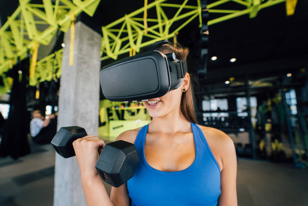 Vue de portrait de la femme portant des lunettes de la réalité virtuelle faisant de l'exercice avec des haltères au gymnase. - Photo, image
