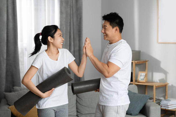 Positieve Aziatische middelbare leeftijd man en jonge vrouw in sportkleding met yoga matten geven elkaar high five en glimlachen, vieren mooie ochtend workout, huis interieur, kopieerruimte - Foto, afbeelding