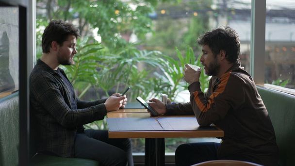 Двоє чоловіків сидять у кав'ярні, дивлячись на мобільні телефони окремо кожен у своїх технологічних бульбашках
 - Фото, зображення