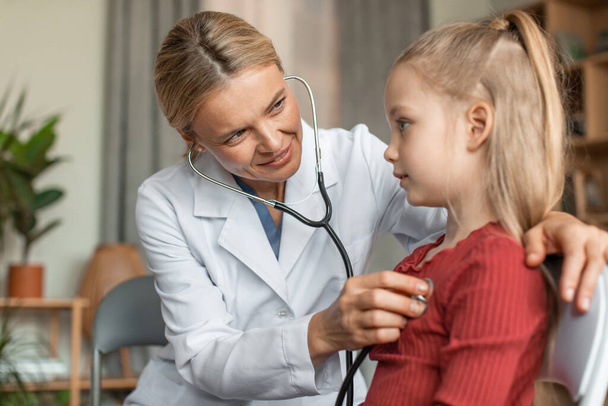 Caring female cardiologist holding stethoscope and listening to girl kid patient at consultation. Woman doctor examining little child client at meeting in hospital. Healthcare concept - Photo, Image