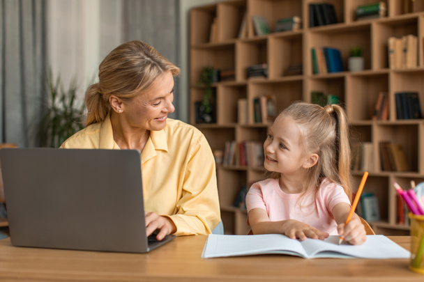 Happy mother and her little daughter spending time together, cute preschool girl drawing at table while her mom working remotely on laptop computer at home - Fotó, kép