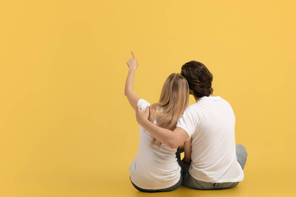 Joyeux couple romantique de jeunes caucasiens en t-shirts blancs étreignant, montrer doigt à l'espace vide, s'asseoir sur le sol, isolé sur fond jaune, dos. Date, grande annonce et offre, vente, amour et relation - Photo, image