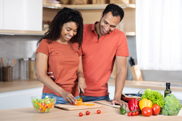 Preparing lunch for family at home. Glad millennial black husband hugging lady, guy cuts organic vegetables at salad in kitchen interior. Support, cooking together, household chores during covid-19 - Foto, imagen