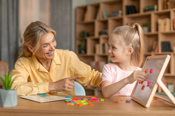 Excited woman speech therapist teaching adorable little girl alphabet, making words from plastic letters on whiteboard. Speech-language therapy for children concept - 写真・画像