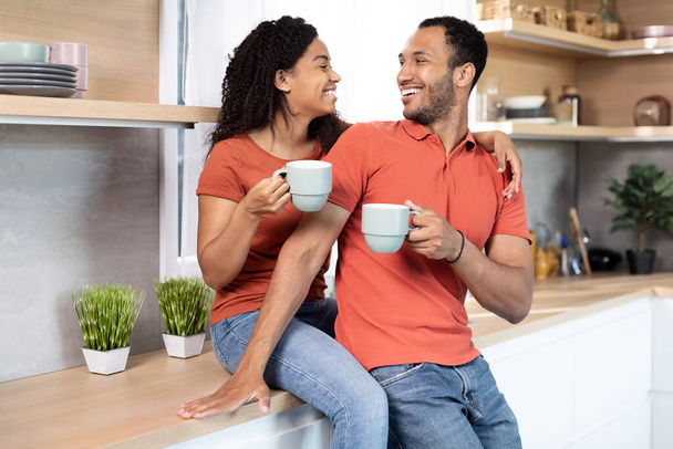 Smiling millennial black woman hugging man, cheers cups with coffee, enjoy free time in kitchen interior. Breakfast together, holiday celebration at home, rest and relax at weekend and good morning - Фото, изображение