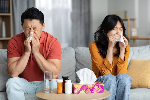 Sick asian husband and wife in casual outfit sitting on sofa next to tea table full of medicine and sneezing nose, using napkins, copy space. Cold, flu, coronavirus concept - Photo, Image